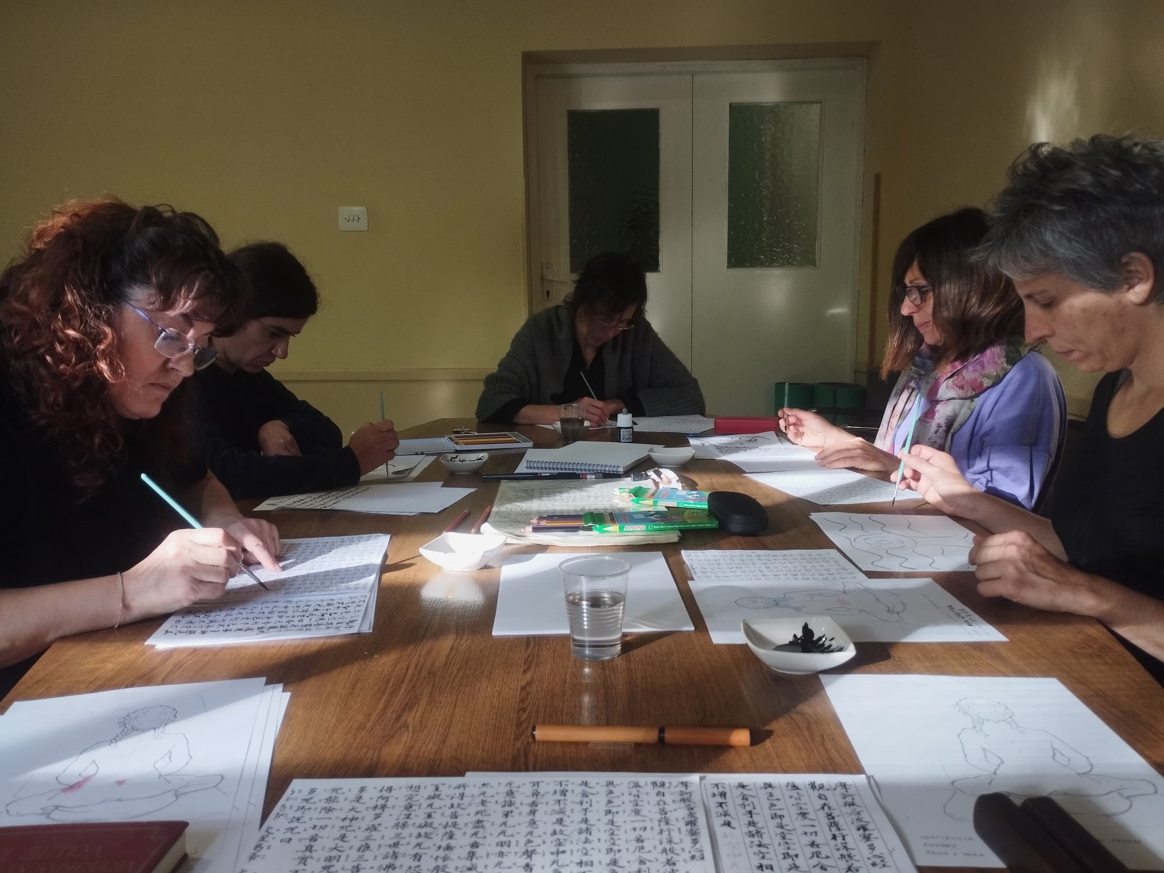 Varias personas practican caligrafía alrededor de una mesa, con pinceles y hojas de papel.
